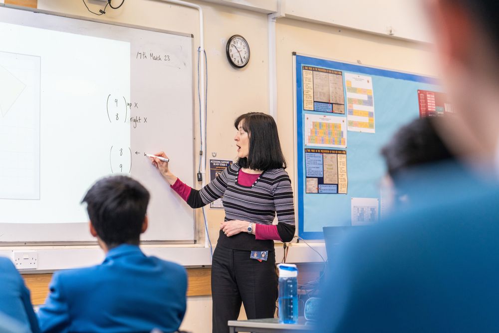 Teacher at whiteboard writing equations with pupils watching.